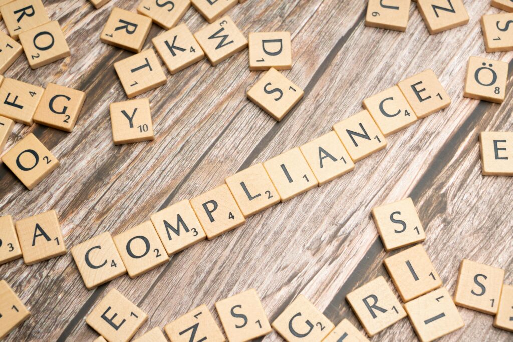 Scrabble tiles spelling "COMPLIANCE" on a wooden surface, surrounded by scattered letter tiles.
